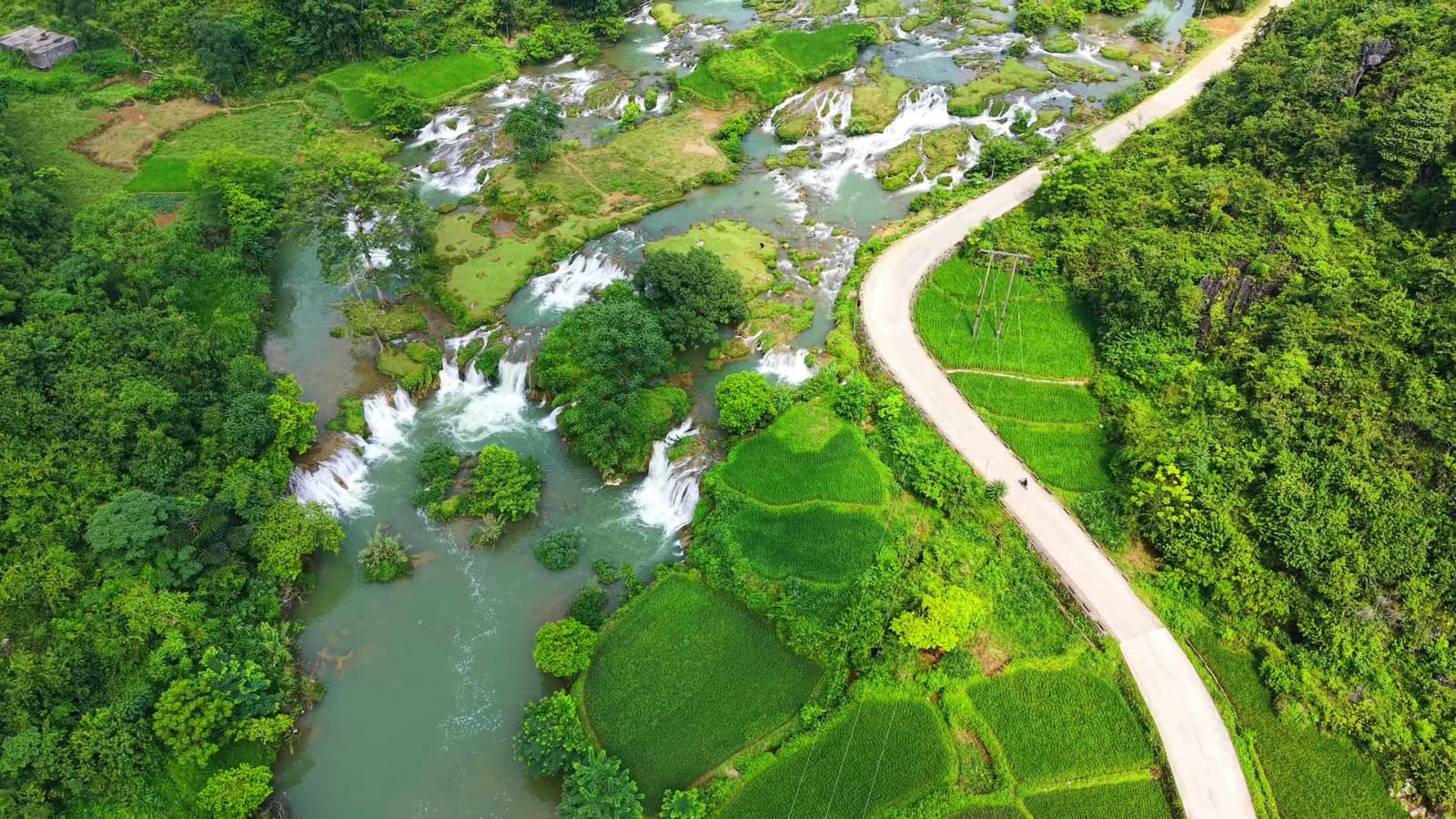 Dòng suối với nhiều thác nhỏ chảy qua thung lũng xanh, bên cạnh con đường uốn cong