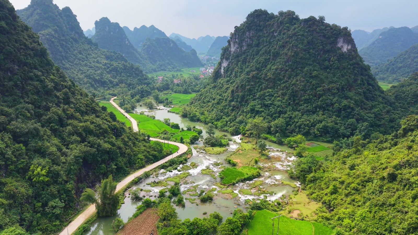 Thung lũng núi đá vôi với dòng suối, ruộng xanh và con đường uốn quanh chân núi